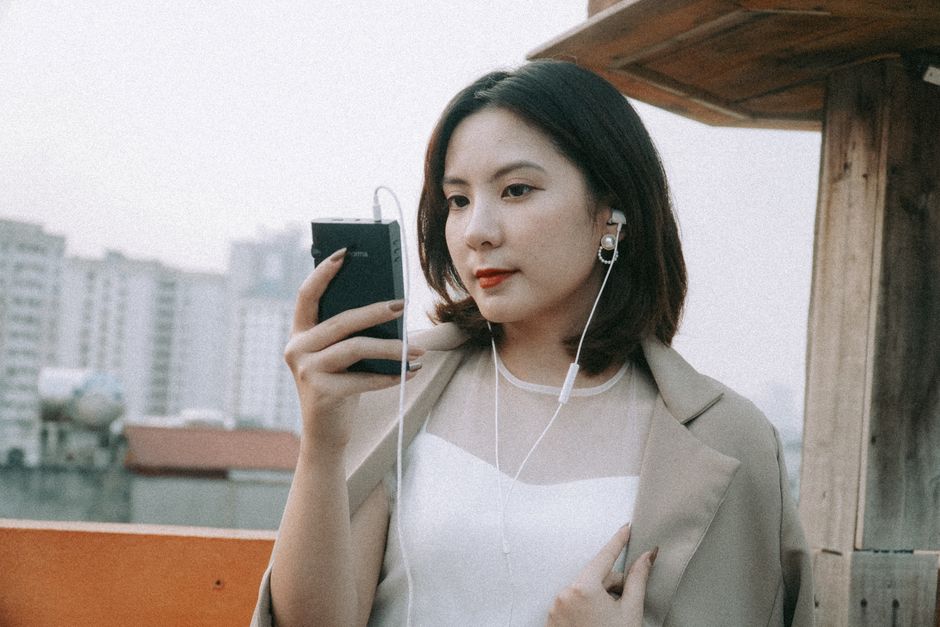 Young woman in stylish attire listens to music using her smartphone on a rooftop with cityscape background.