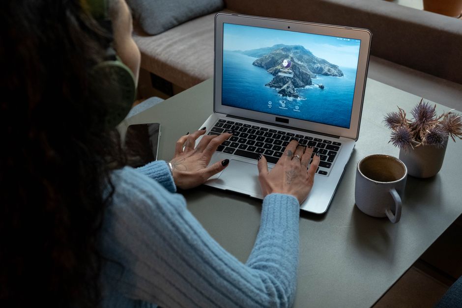 A woman wearing headphones works on a laptop at a cozy home office with a cup of coffee nearby.